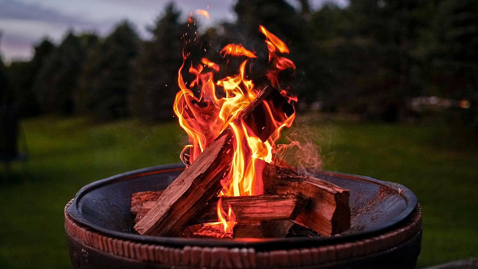 Landscaping Toronto - A fire pit with burning logs and rising flames in an outdoor patio setting at dusk, surrounded by trees.