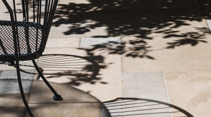 Landscaping Toronto - Patterned shadows of leaves fall onto a natural stone patio floor, intersecting with the shadow of a metal chair in bright sunlight.