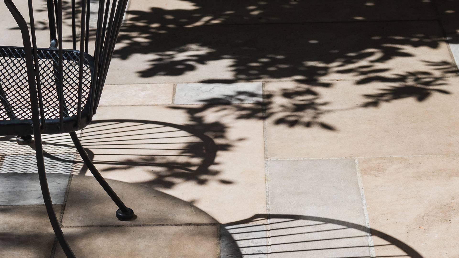Landscaping Toronto - Patterned shadows of leaves fall onto a natural stone patio floor, intersecting with the shadow of a metal chair in bright sunlight.